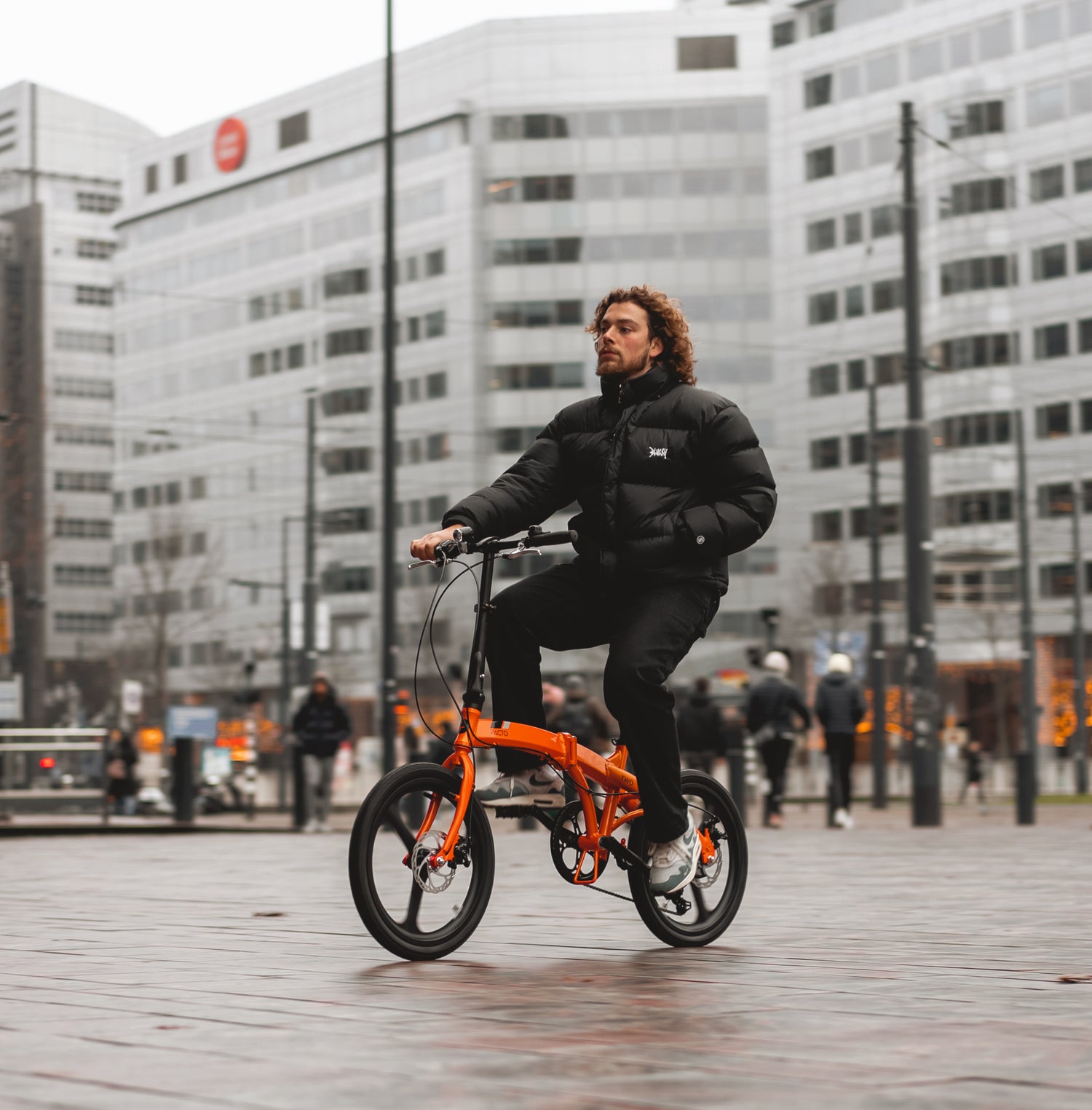 Man riding a bright orange PACTO Two folding bike in a modern city square, highlighting its compact urban commuter design.
