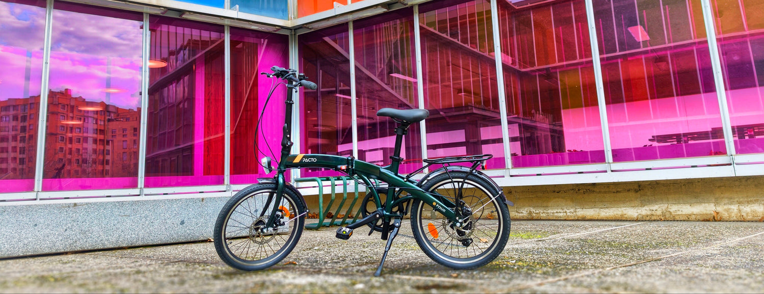 Green PACTO FOUR S folding bike with Shimano gears parked in front of colorful modern building
