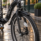 Nederlands:
Detail van het voorwiel van de zwarte PACTO SEVEN vouwfiets met koplamp, voorrem en spatbord op een natte stadsstraat.

Engels:
Close-up of the front wheel of the black PACTO SEVEN folding bike with headlight, front brake and mudguard on a wet city street.
