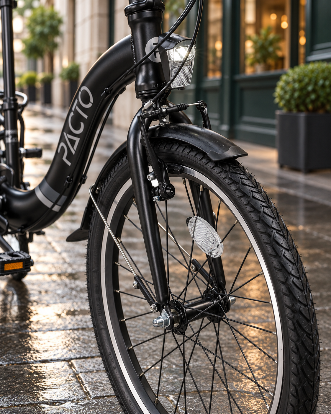 Nederlands:
Detail van het voorwiel van de zwarte PACTO SEVEN vouwfiets met koplamp, voorrem en spatbord op een natte stadsstraat.

Engels:
Close-up of the front wheel of the black PACTO SEVEN folding bike with headlight, front brake and mudguard on a wet city street.