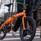 PACTO Two folding bike in bright orange being walked across a city crosswalk, showcasing its compact urban design.