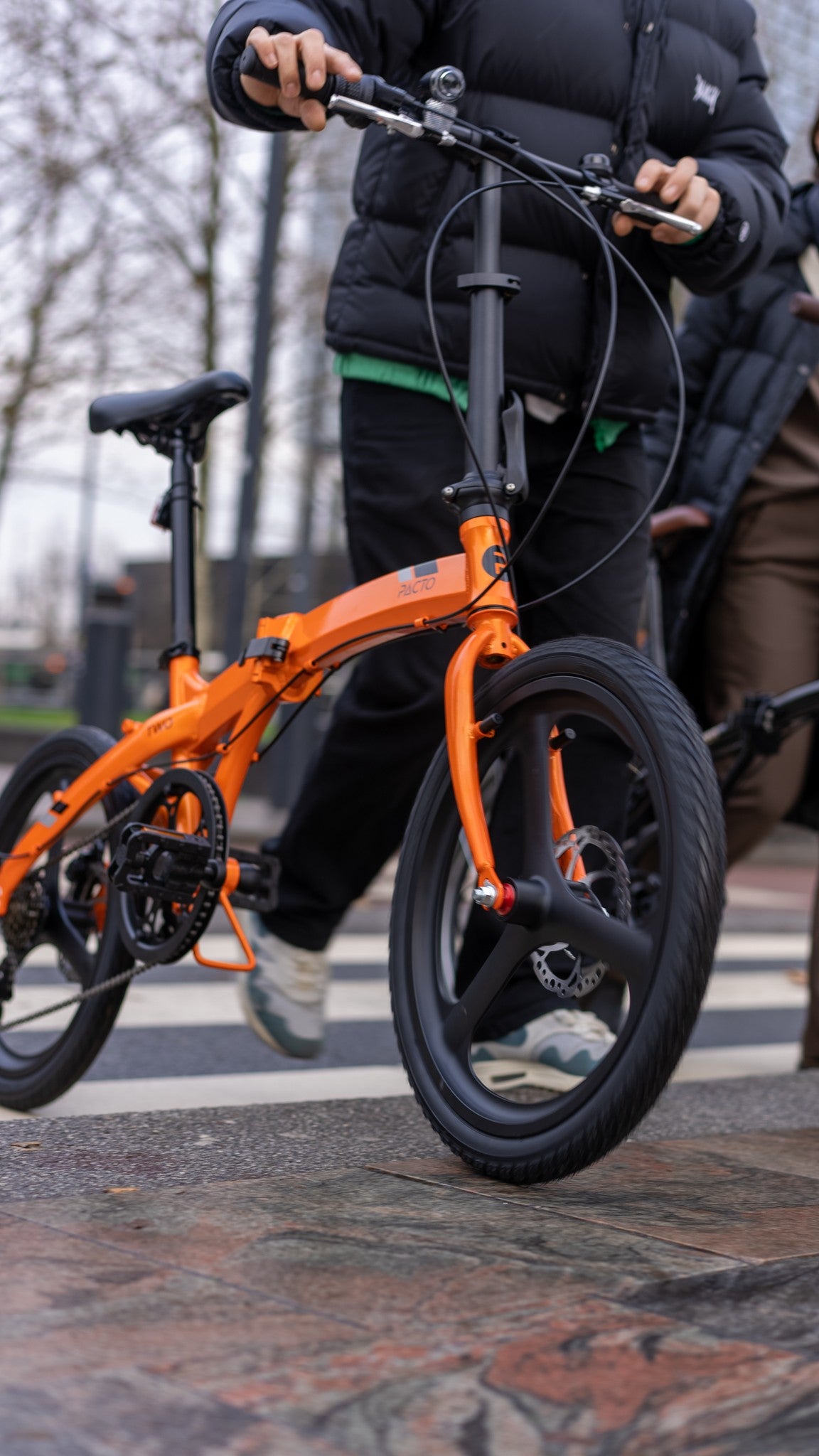 PACTO Two folding bike in bright orange being walked across a city crosswalk, showcasing its compact urban design.